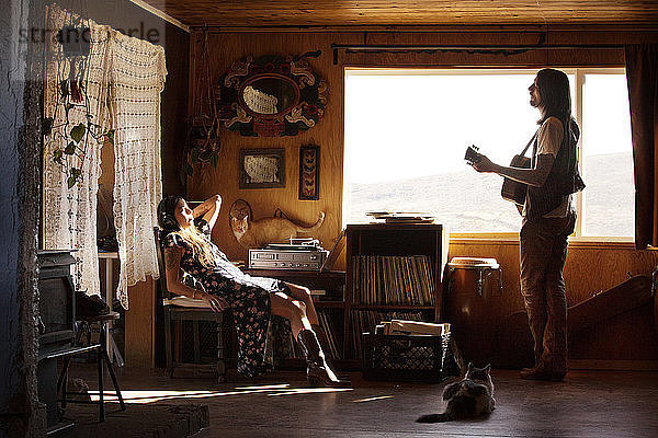 Mann spielt Gitarre  während sich die Frau zu Hause auf einem Stuhl entspannt