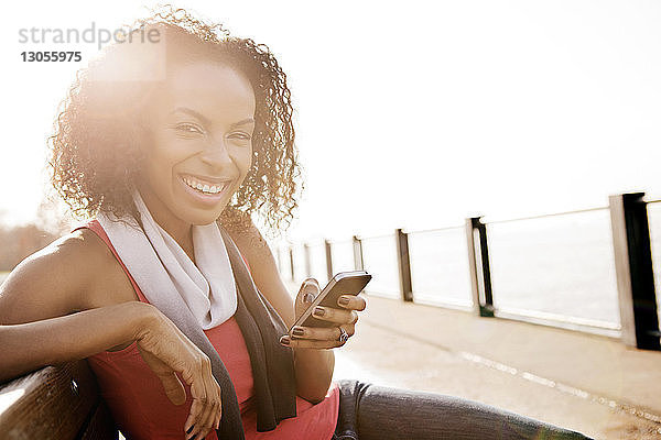 Porträt einer glücklichen Frau  die an einem sonnigen Tag auf einer Bank sitzend ihr Handy in der Hand hält