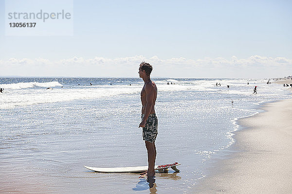 Mann schaut weg  während er mit einem Surfbrett am Ufer steht