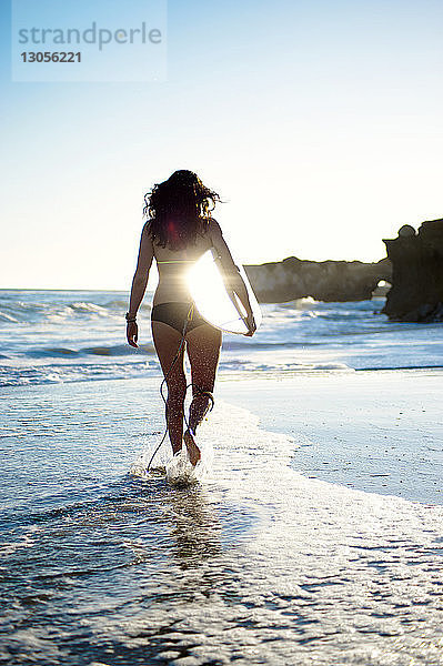 Rückansicht einer Frau mit Surfbrett beim Strandspaziergang am Ufer