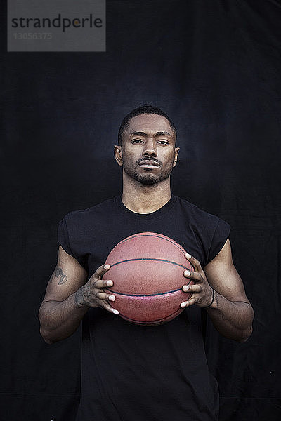 Porträt eines Sportlers  der Basketball in der Hand hält  während er gegen eine schwarze Wand steht