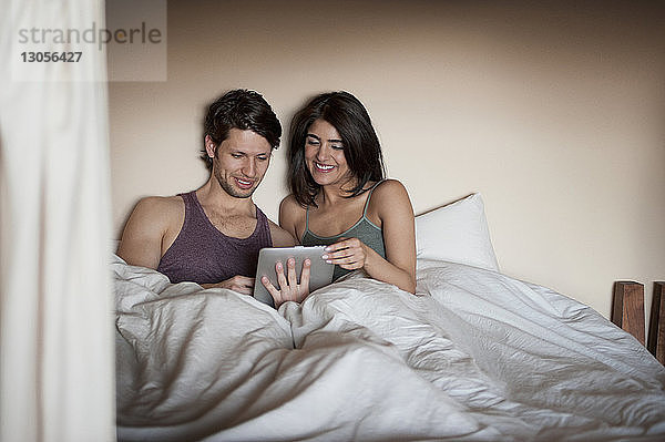 Fröhliches junges Paar betrachtet digitales Tablett auf dem Bett