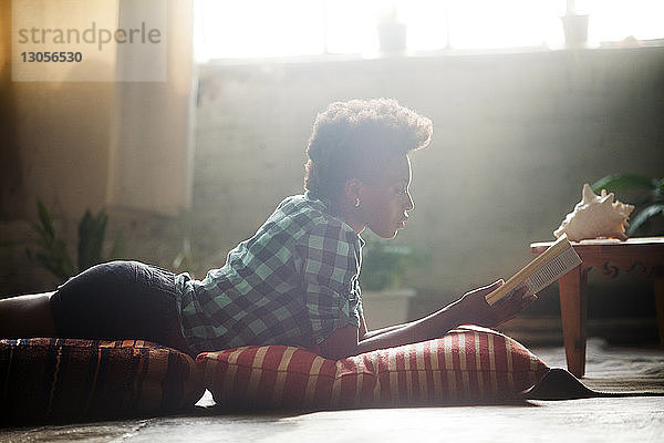 Seitenansicht einer Frau  die im Wohnzimmer auf Kissen liegend ein Buch liest