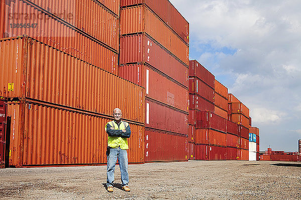 Porträt eines selbstbewussten Mannes mit verschränkten Armen  der gegen Frachtcontainer steht