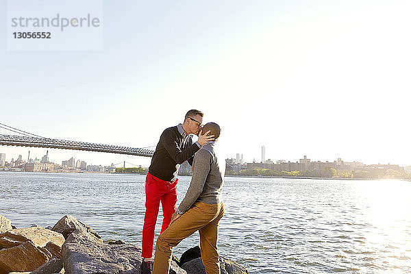 Mann küsst seinen Freund  während er an einem sonnigen Tag auf Felsen am East River steht