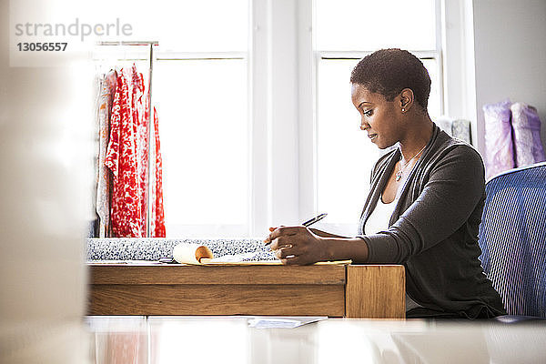 Seitenansicht einer Modedesignerin  die am Schreibtisch gegen das Fenster arbeitet