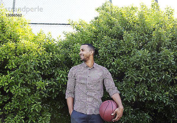 Glücklicher junger Mann hält Basketball  während er gegen Pflanzen steht