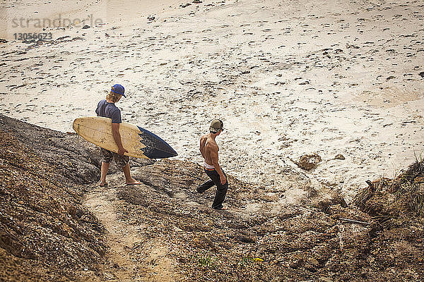 Mann trägt Surfbrett  während er mit einem Freund am Strand spazieren geht
