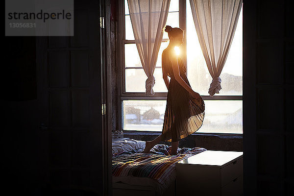 Seitenansicht einer Frau in einem Kleid  die auf dem Bett am Fenster steht und durch die Tür gesehen wird