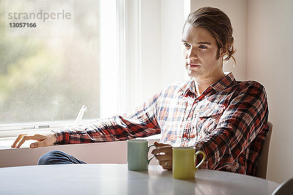 Nachdenklicher Mann hält Kaffeetasse  während er zu Hause am Fenster sitzt