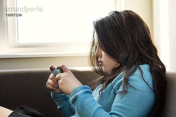 Seitenansicht eines Mädchens  das ein Mobiltelefon benutzt  während es sich zu Hause auf dem Sofa entspannt