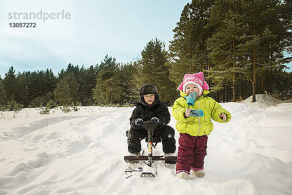 Junge sitzt auf Schlitten  während die Schwester auf schneebedecktem Feld läuft