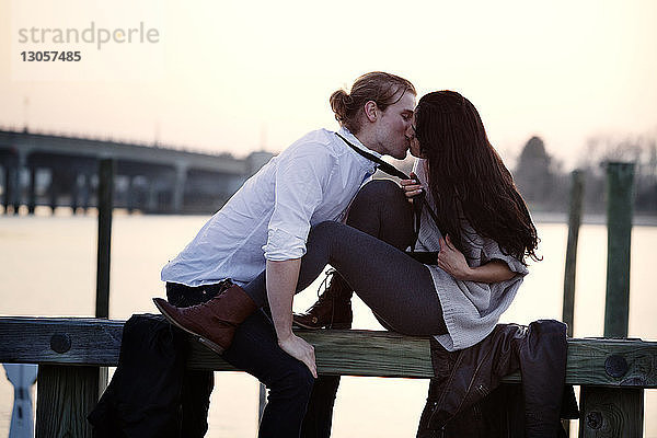 Romantisches Paar küsst sich  während es auf einem Geländer gegen den Himmel sitzt