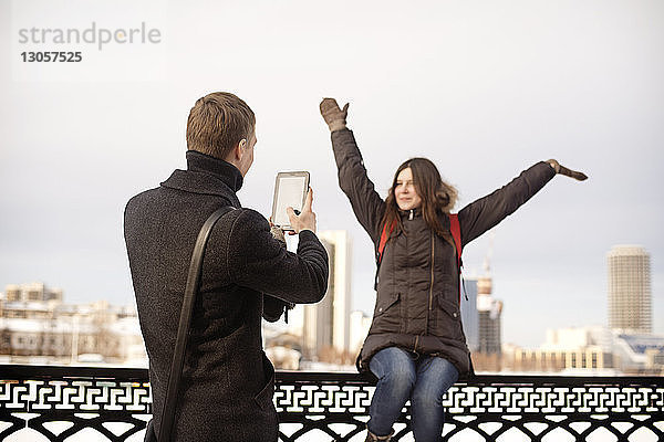 Mann fotografiert glückliche Freundin  die auf einem Geländer im Park sitzt