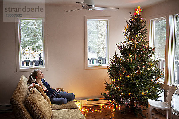 Frau schaut auf den Weihnachtsbaum  während sie auf dem Sofa sitzt