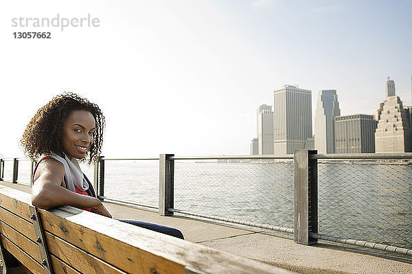 Porträt einer lächelnden jungen Frau auf einer Bank am East River in der Stadt vor klarem Himmel