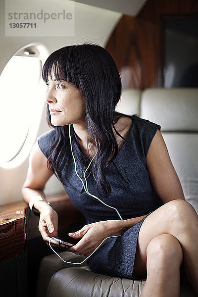 Geschäftsfrau hört Musik  während sie im Firmenjet durchs Fenster schaut