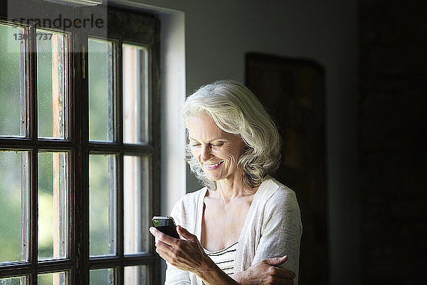 Glückliche ältere Frau telefoniert  während sie zu Hause am Fenster steht