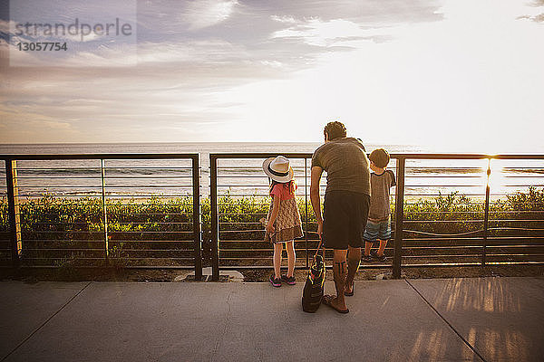 Rückansicht einer Familie  die bei Sonnenuntergang am Geländer steht und aufs Meer schaut