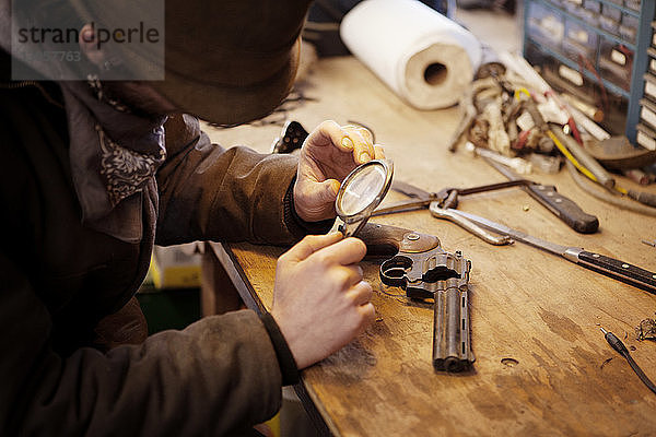 Hochwinkelansicht eines Mannes  der die Pistole mit Lupe bei Tisch in der Werkstatt untersucht