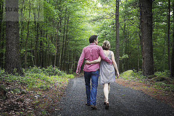 Rückansicht eines Paares mit umarmenden Armen  das auf einem Schotterweg im Wald geht