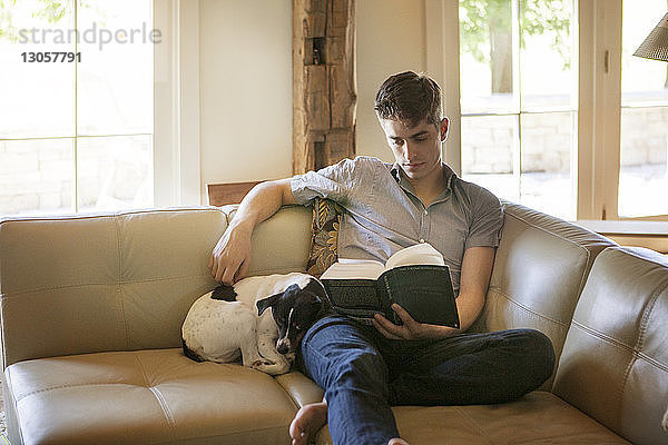 Mann liest Buch  während er zu Hause mit Hund auf dem Sofa sitzt