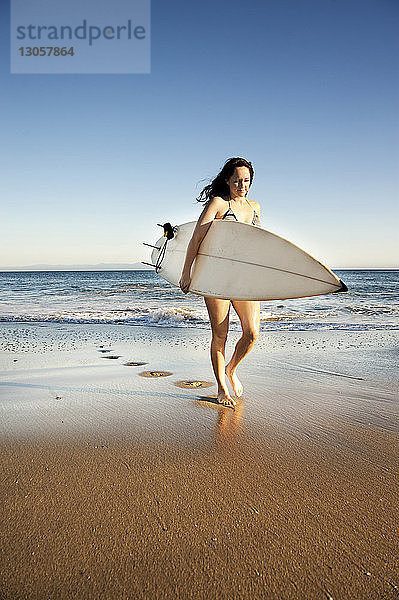 Frau geht am Strand mit Surfbrett am Ufer spazieren