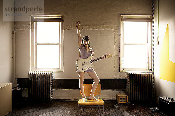 Glückliche Frau spielt Gitarre  während sie zu Hause auf einem Stuhl steht