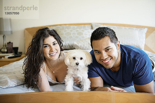 Porträt eines glücklichen Paares mit Hund  das zu Hause auf dem Bett liegt