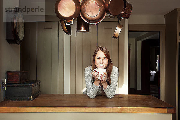 Porträt einer glücklichen Frau  die eine Kaffeetasse hält  während sie sich zu Hause auf eine Holztheke stützt