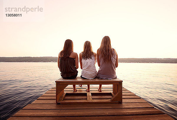 Rückansicht von Freunden  die bei Sonnenuntergang auf einem Steg über dem See vor klarem Himmel sitzen