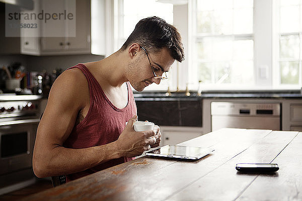 Mann mit Kaffeetasse schaut auf Tablet-Computer  während er in der Küche am Tisch sitzt