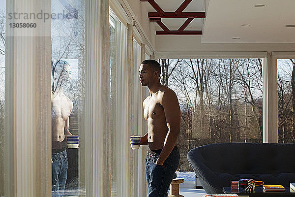 Mann ohne Hemd hält Becher  während er zu Hause durchs Fenster schaut