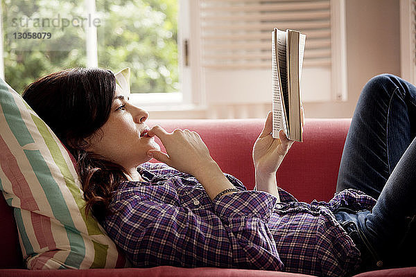 Nachdenkliche Frau liest Buch  während sie zu Hause auf dem Sofa liegt