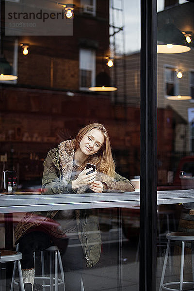 Lächelnde Frau mit Smartphone im Café durchs Fenster gesehen