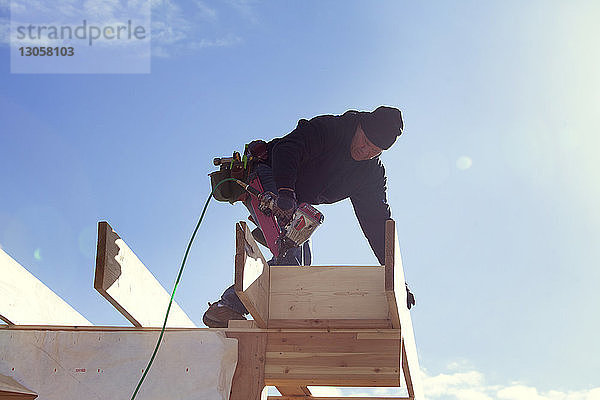 Niedrigwinkelansicht eines Arbeiters mit Bohrmaschine  der auf einem Dachbalken gegen den Himmel steht