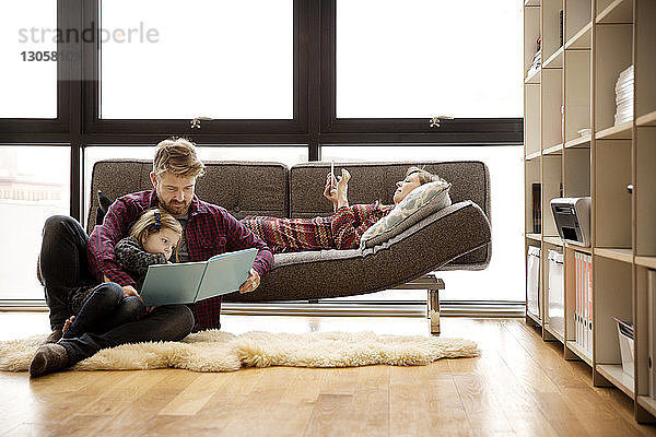 Vater und Tochter lesen ein Buch von einer Frau  die sich zu Hause auf dem Sofa entspannt