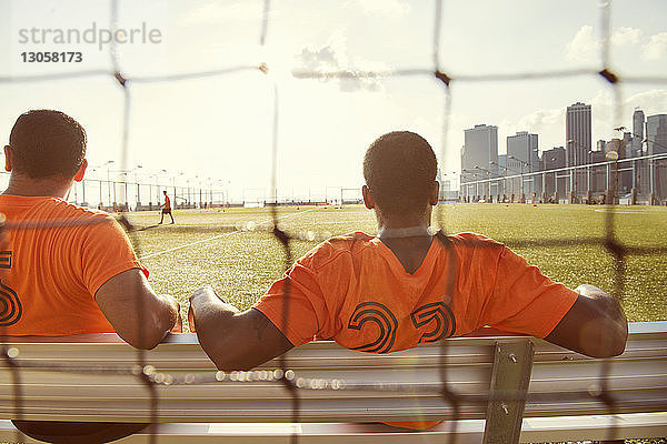 Rückansicht von Fussballspielern  die auf dem Spielfeld sitzen  durch ein Netz an einem sonnigen Tag gesehen