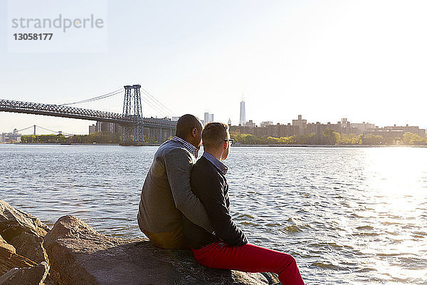 Seitenansicht von schwulen Männern  die auf Felsen sitzen  während sie an einem sonnigen Tag auf den East River blicken
