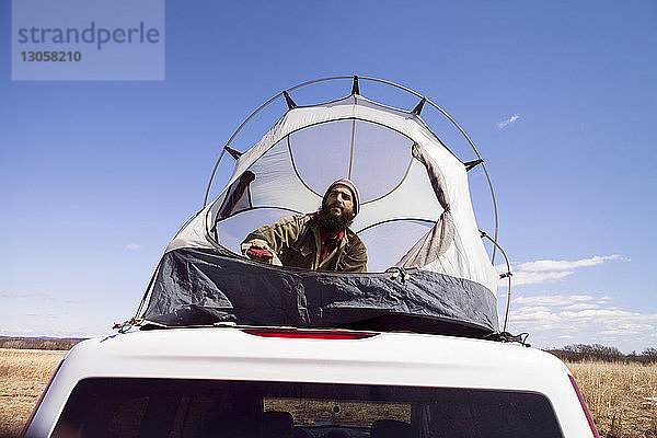 Niedrigwinkelansicht eines nachdenklichen Mannes  der im Zelt auf dem Autodach vor blauem Himmel sitzt