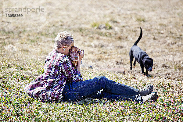 Seitenansicht eines Jungen  der mit Hunden auf dem Feld sitzt