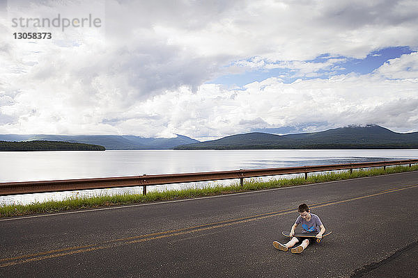 Junge mit Skateboard sitzt auf Straße gegen bewölkten Himmel