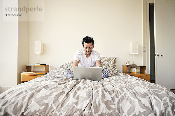 Mann benutzt Laptop  während er zu Hause auf dem Bett sitzt