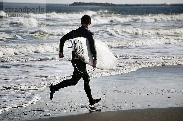 Rückansicht eines Surfers  der ein Surfbrett trägt  während er am Strand an Land läuft