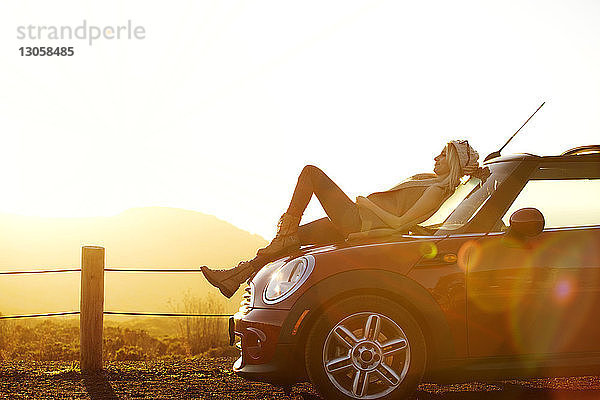 Seitenansicht einer Frau  die sich auf dem Auto auf dem Feld gegen den Himmel entspannt