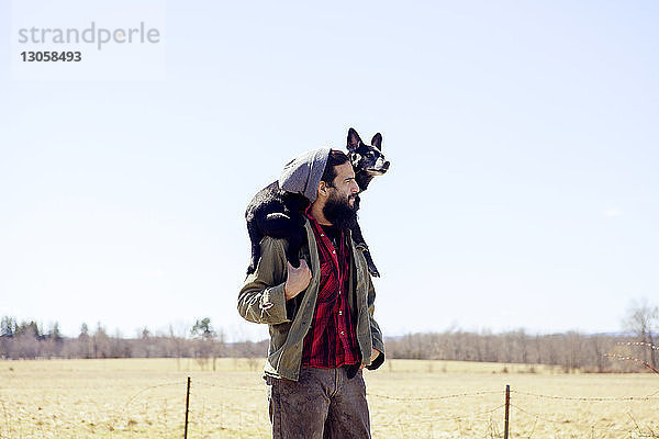Nachdenklicher Mann trägt Hund auf den Schultern  während er auf dem Feld gegen den klaren Himmel steht