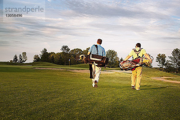 Rückansicht von männlichen Golfern  die bei Sonnenuntergang auf dem Spielfeld Taschen tragen