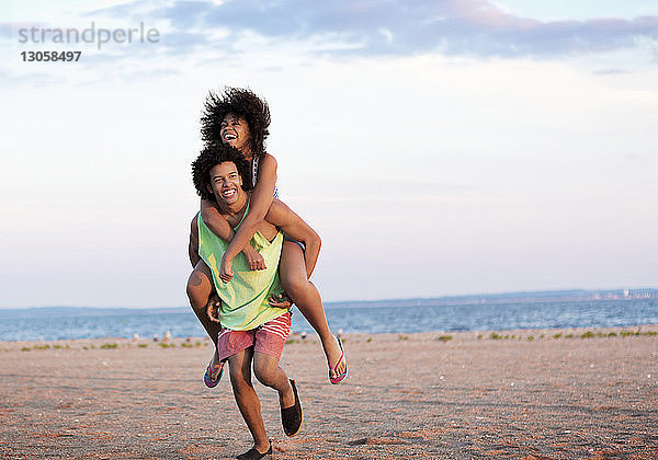 Mann nimmt Freundin am Strand huckepack gegen den Himmel