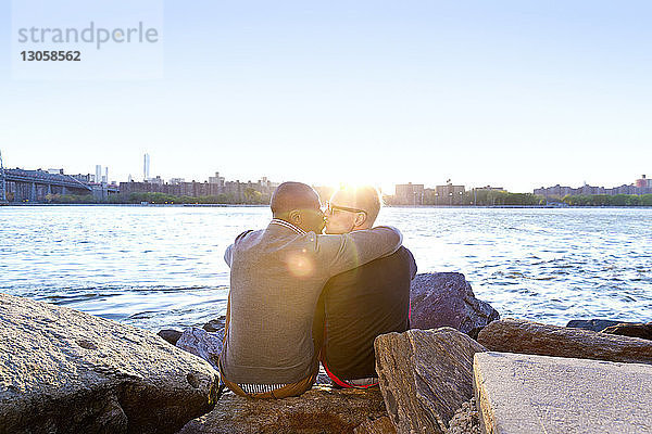 Rückansicht von Freunden  die sich küssen  während sie bei Sonnenuntergang auf Felsen am East River sitzen