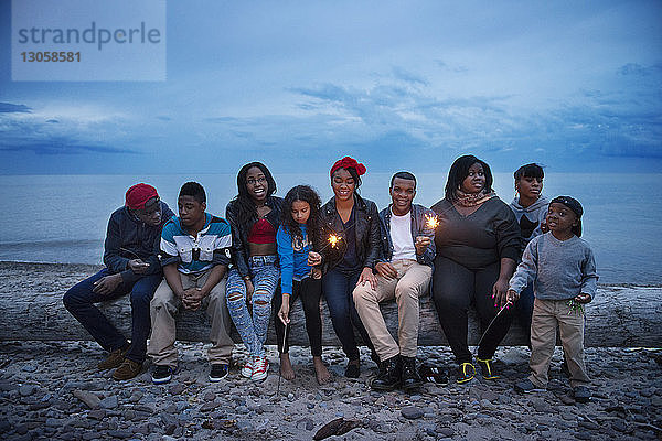Familie und Freunde sitzen auf einem Baumstamm am Strand gegen den Himmel